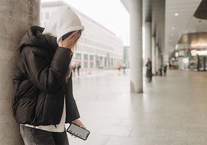 Young person in hoodie with mobile phone in hand covers her face.