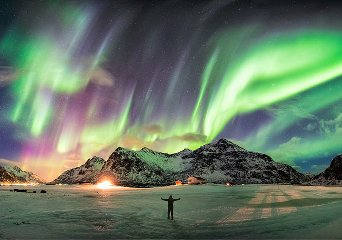A person with outstretched arms on an ice field admires the northern lights in the sky against a mountain backdrop.