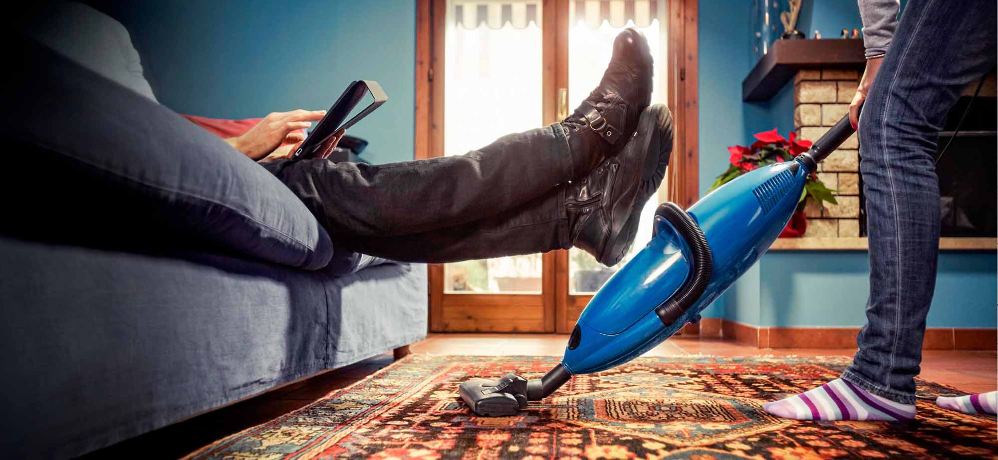 Woman vacuuming carpet under man's feet held up on sofa.